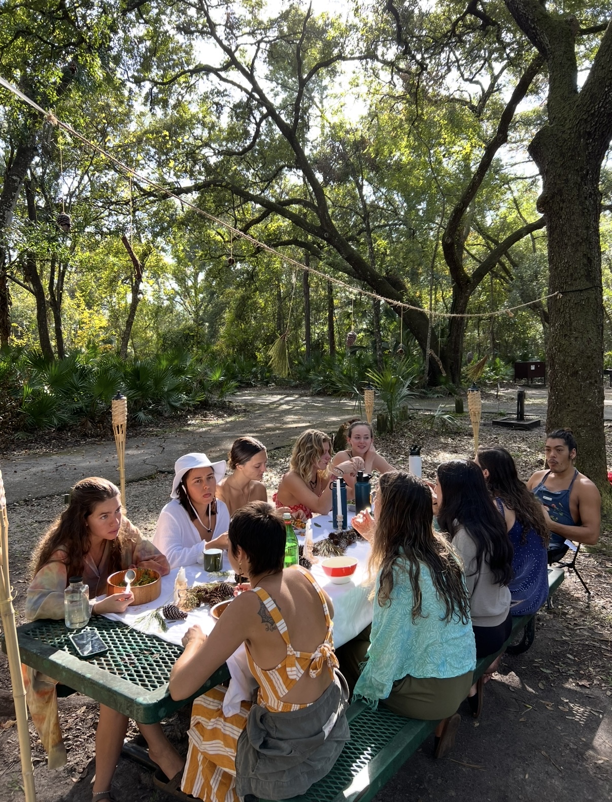 Large group gathered around picnic table in forest, sharing meal together